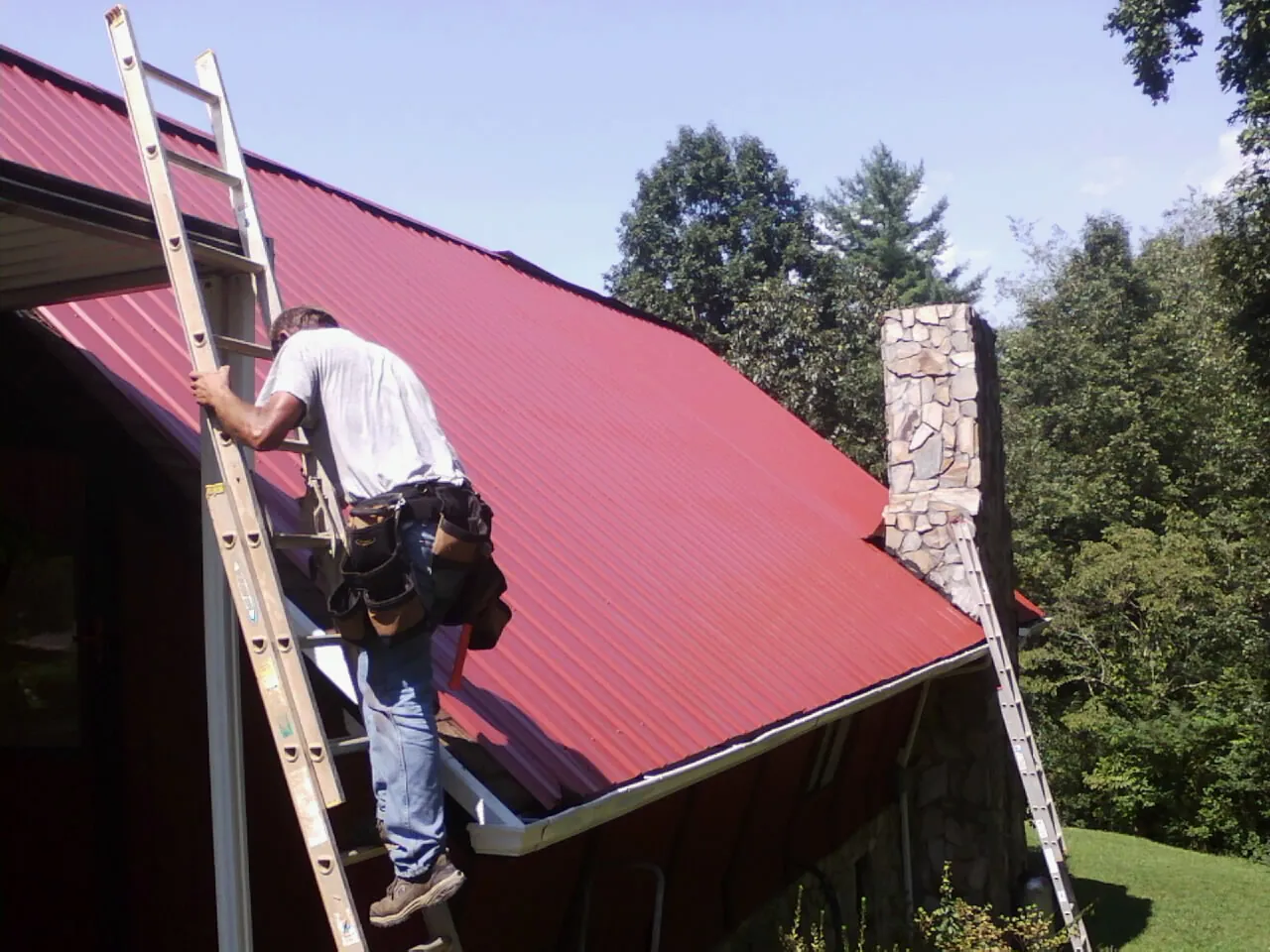 Roofer on a ladder beside a red metal roof and stone chimney — Soffit & Fascia Repair by Roofing Brothers in Rubicon